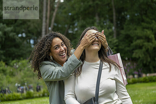 Woman surprises her friend by playfully covering her eyes in a lush park setting  both smiling joyfully.