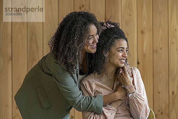 Two women sharing a joyful moment with one embracing the other from behind  both smiling and against a wooden wall background.