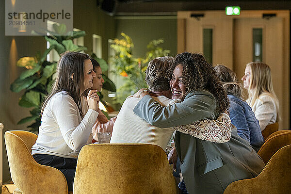 Two women share a warm  genuine hug while others engage in conversation in the cozy ambiance of a modern caf?.