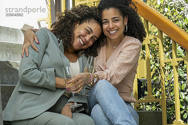 Two women sitting on stairs outdoors  smiling and toasting with champagne glasses.