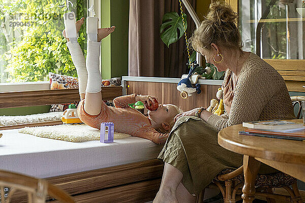 A woman interacts with a happy child lying on a bed with legs raised in cast while toys and a book rest nearby.