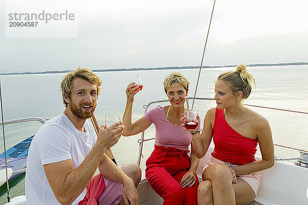 Three friends enjoying drinks and a good time on a boat with a scenic lake in the background during sunset.