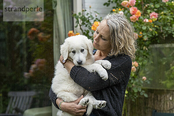 Woman embraces a white puppy in a garden  with flowers in the background.