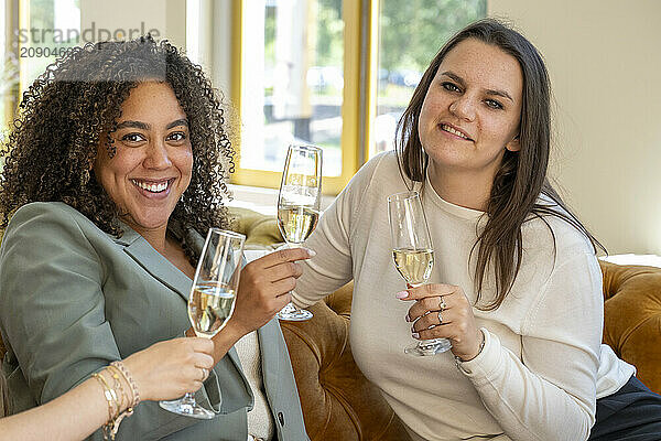 Two women smiling and toasting with glasses of white wine indoors near a window with natural light.