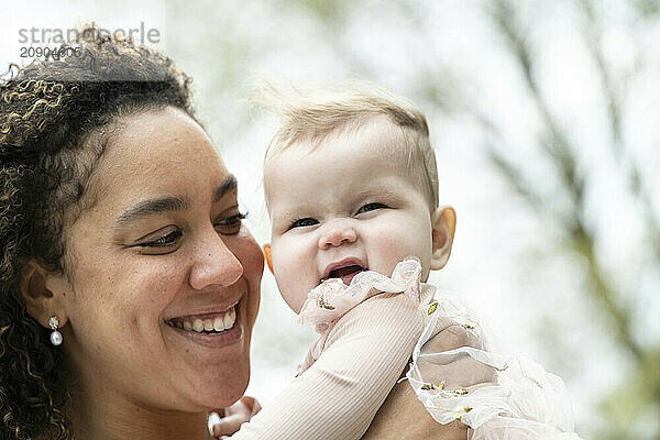 Joyful mother holding her smiling baby girl outdoors on a bright day.