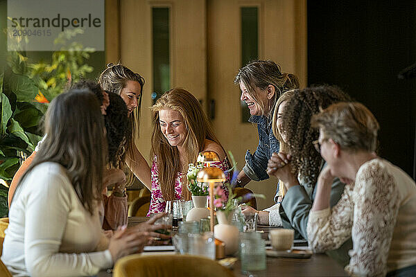 A group of joyful women engaged in conversation around a table adorned with flowers and refreshments in a cozy room with green plants.