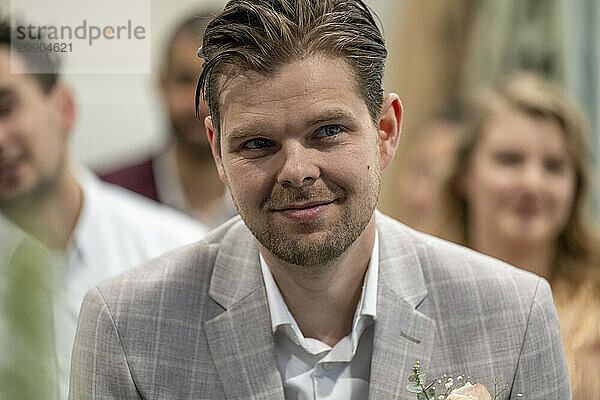 A groom in a gray checkered suit with a boutonniere smiles at a wedding ceremony  with guests blurred in the background.