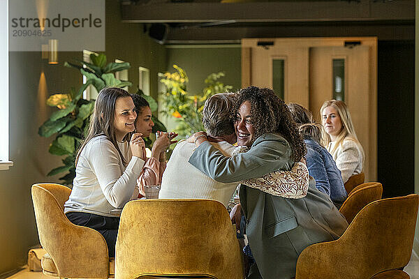 Two women embracing in a warm hug at a cozy gathering  surrounded by others engaged in conversation.