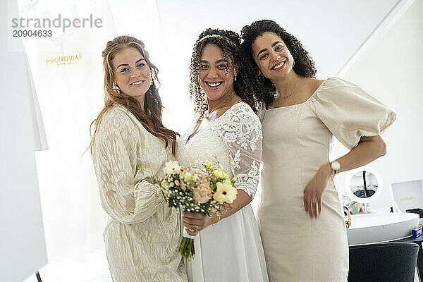 Three women smiling and posing together  one in a wedding dress holding a bouquet  flanked by two others in casual dresses.