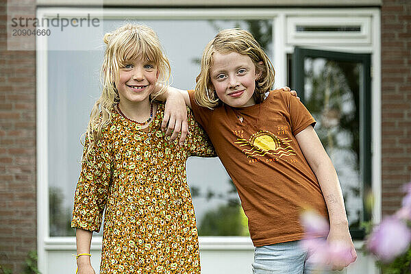 Two smiling girls embracing each other in front of a house with a large window and greenery around.