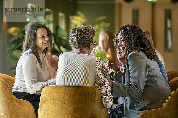 A group of women enjoy a lively conversation while seated on yellow chairs around a table in a cozy room.