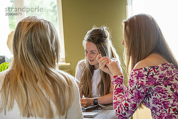 Three women engage in a lively conversation at a dining table with natural light filtering through the window.