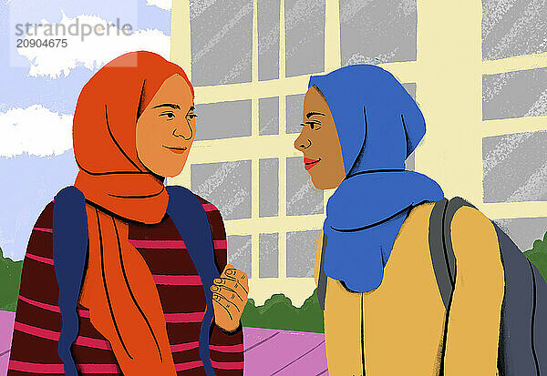 Two smiling women wearing hijabs and backpacks are engaging in a friendly conversation against the backdrop of a building.