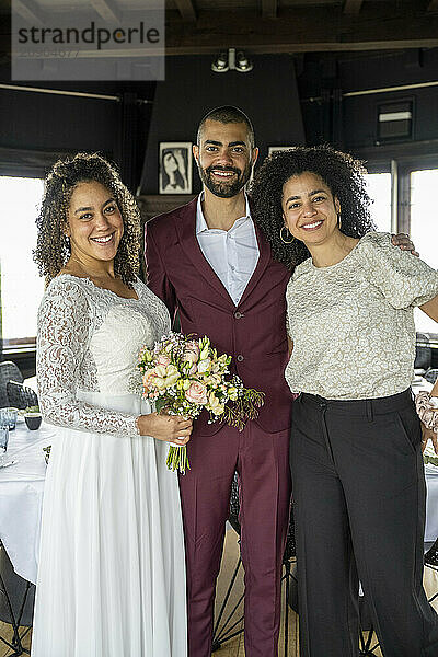 Smiling bride in a white dress holding a bouquet stands beside a groom in a burgundy suit and a woman in a gray lace top.