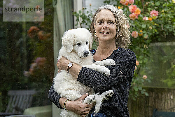 Smiling woman holding a fluffy white puppy in a garden with blooming flowers