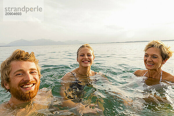 Three people are joyfully swimming in an open water setting with mountains in the background at sunset.