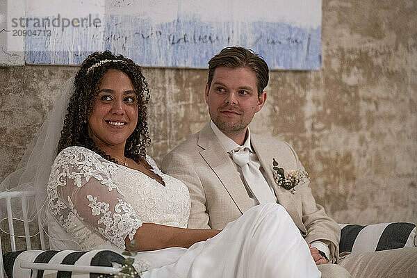 Bride and groom smiling while seated at their wedding  with the bride in a lace dress and the groom in a beige suit.