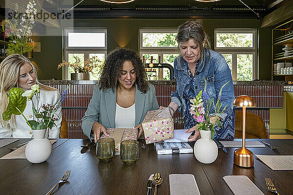 Three women are smiling and examining a gift box at a beautifully set dining table with modern decor and flowers.