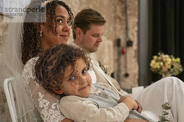 A bride in a lace wedding dress lovingly gazes aside while a young boy rests his head on her lap  with a man in the background.