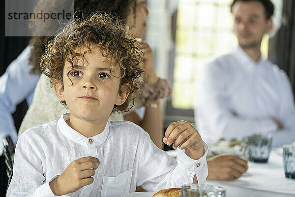 Curly-haired child in a white shirt sitting at a dining table with adults blurred in the background.