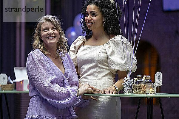 Two smiling women holding hands at a celebration table with decorative balloons and favors.