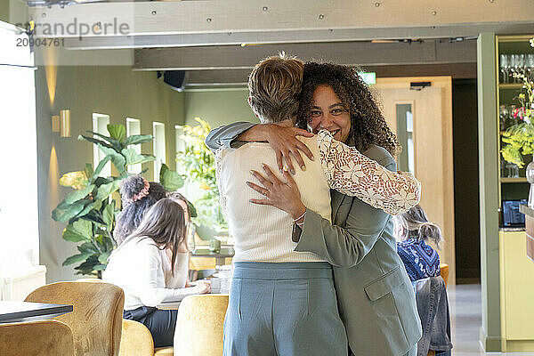 Two women are warmly embracing inside a bright cafe with other patrons visible in the background.