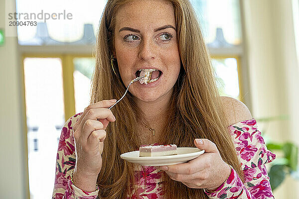 Woman with long red hair enjoys a slice of cake with a fork in a bright room.