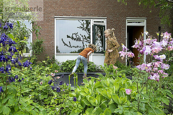 Child jumping on a trampoline in a garden with a woman watching  surrounded by lush green plants and flowers.