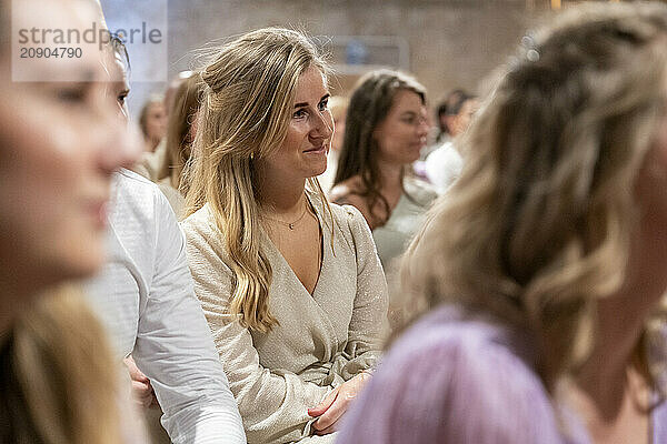 Smiling woman in a beige sweater attentively sitting in an audience with other attendees in soft focus.