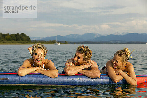 Three friends enjoy a sunny day on a paddleboard in a calm lake with mountains in the distance.