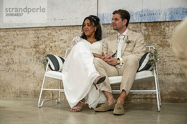 A bride and groom sit closely together on a white bench  smiling contentedly amid their wedding celebrations.