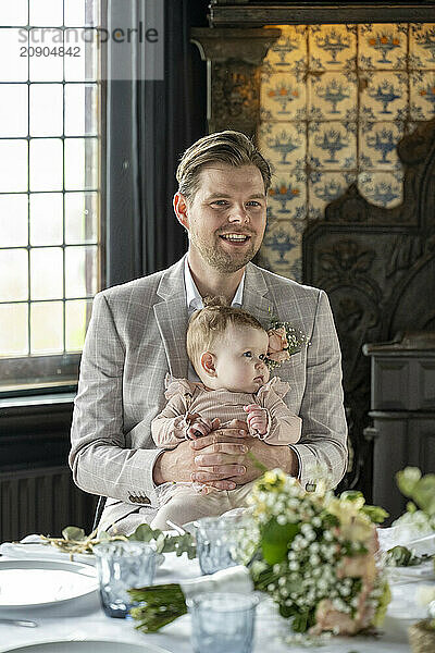 A well-dressed man smiles as he holds a baby in a formal setting with vintage decor.