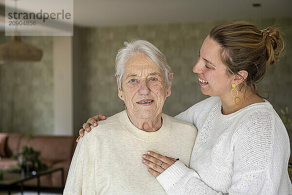 Elderly woman with a heartwarming smile embraced by a joyful young woman in a cozy home interior.