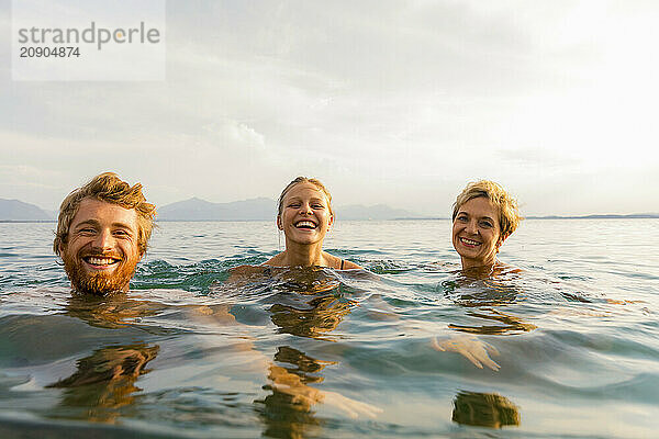 Three people are smiling and swimming in an open water setting with mountains in the background under a clear sky.