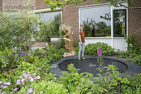 Girl in a floral dress joyfully jumping on a garden trampoline while another girl in orange looks on  with lush greenery and a house in the background.