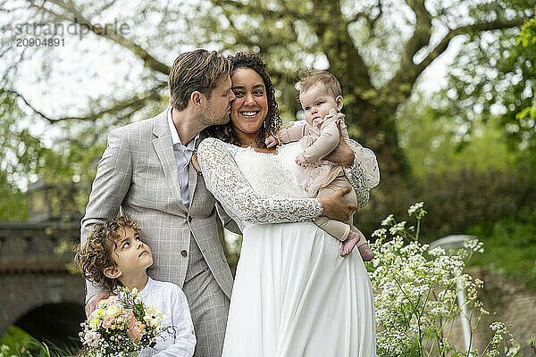 Happy family dressed in formal attire posing in a lush park  with a father kissing the mother on the cheek while she holds a baby  and a young child holding a floral bouquet standing beside them.