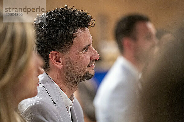 Side profile of a smiling man with curly hair attending an event  with other attendees blurred in the foreground and background.