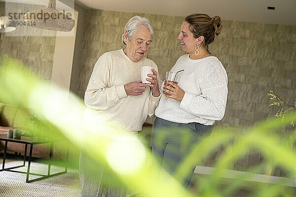 Elderly woman and young woman smiling and holding cups in a cozy home environment.