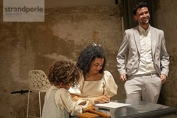 A smiling groom stands as a bride signs a document at a table with a young child watching attentively in a room with rustic decor.