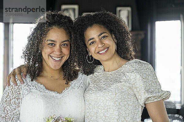 Two smiling women with curly hair embracing each other in a room with natural light.