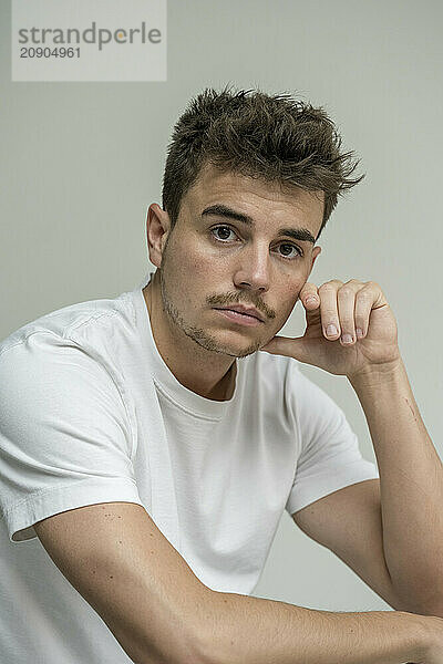 A pensive young man with stubble sits with his chin resting on his hand against a plain background.