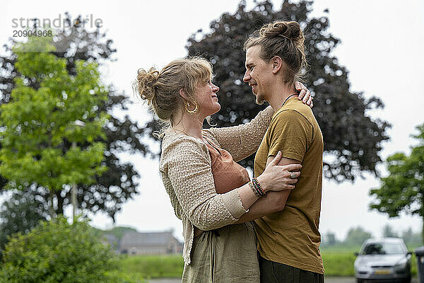 A smiling woman and man embrace in a rural setting with trees in the background.