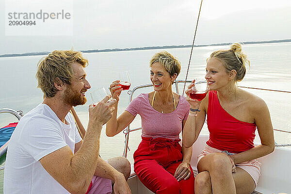 Three friends enjoying drinks and conversation on the deck of a sailboat with a serene water backdrop at dusk.