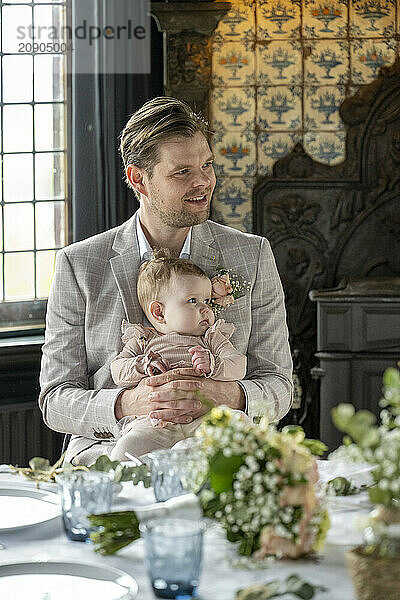 Father in a suit holding his baby at a formal event with decorative floral arrangements on the table.