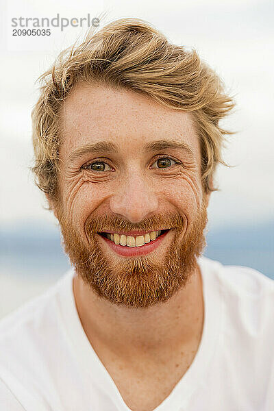 Smiling man with a beard and messy hair against a blurred background.