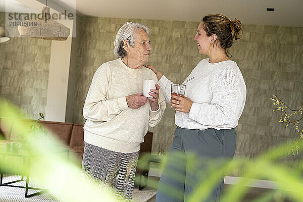 Elderly woman and younger adult female sharing a conversation and drinks in a cozy living room.