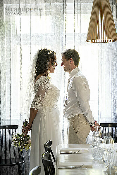 Bride in a lace dress and groom in a white shirt standing close to each other near a table set for a celebration  with sheer curtains diffusing the sunlight behind them.