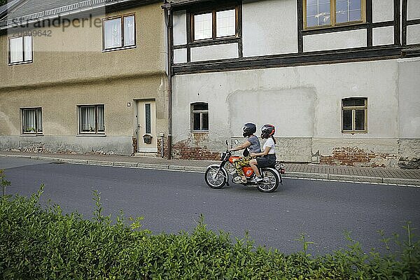 Symbolfoto zum Thema Jugend im Osten. Ein Junge und ein Mädchen fahren auf einem Simson Moped an alten Häusern vorbei ueder eine Dorfstraße Ziegenrück  20.07.2024