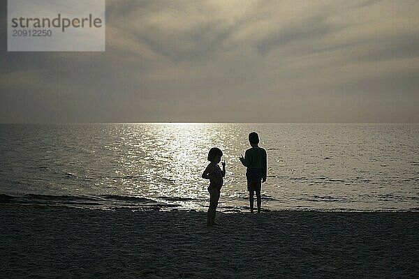 Geschwister stehen im Gegenlicht der untergehenden Sonne am Strand. Born  01.08.2024