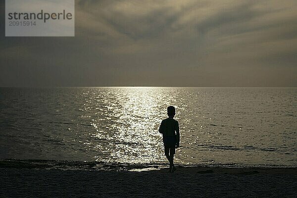 Ein Kind steht alleine im Gegenlicht der untergehenden Sonne am Strand. Born  01.08.2024